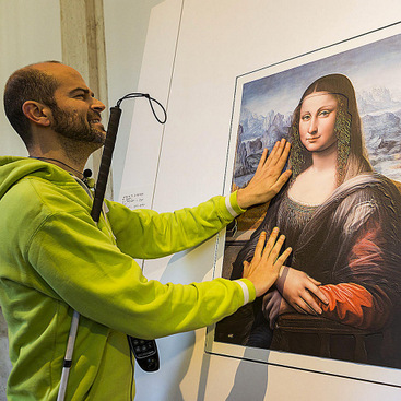 A person with a cane, black glasses, and a device on a waist is touching a tactile artwork in the museum.